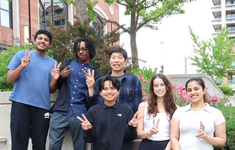 Six SLIDE Studio lab members posing with peace signs outdoors in front of greenery and roses